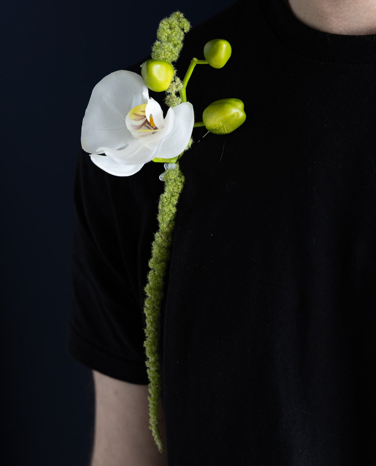 A white orchid flower boutonniere with green buds and a textured green stem, pinned to the left shoulder of a person wearing a black t-shirt against a dark background.