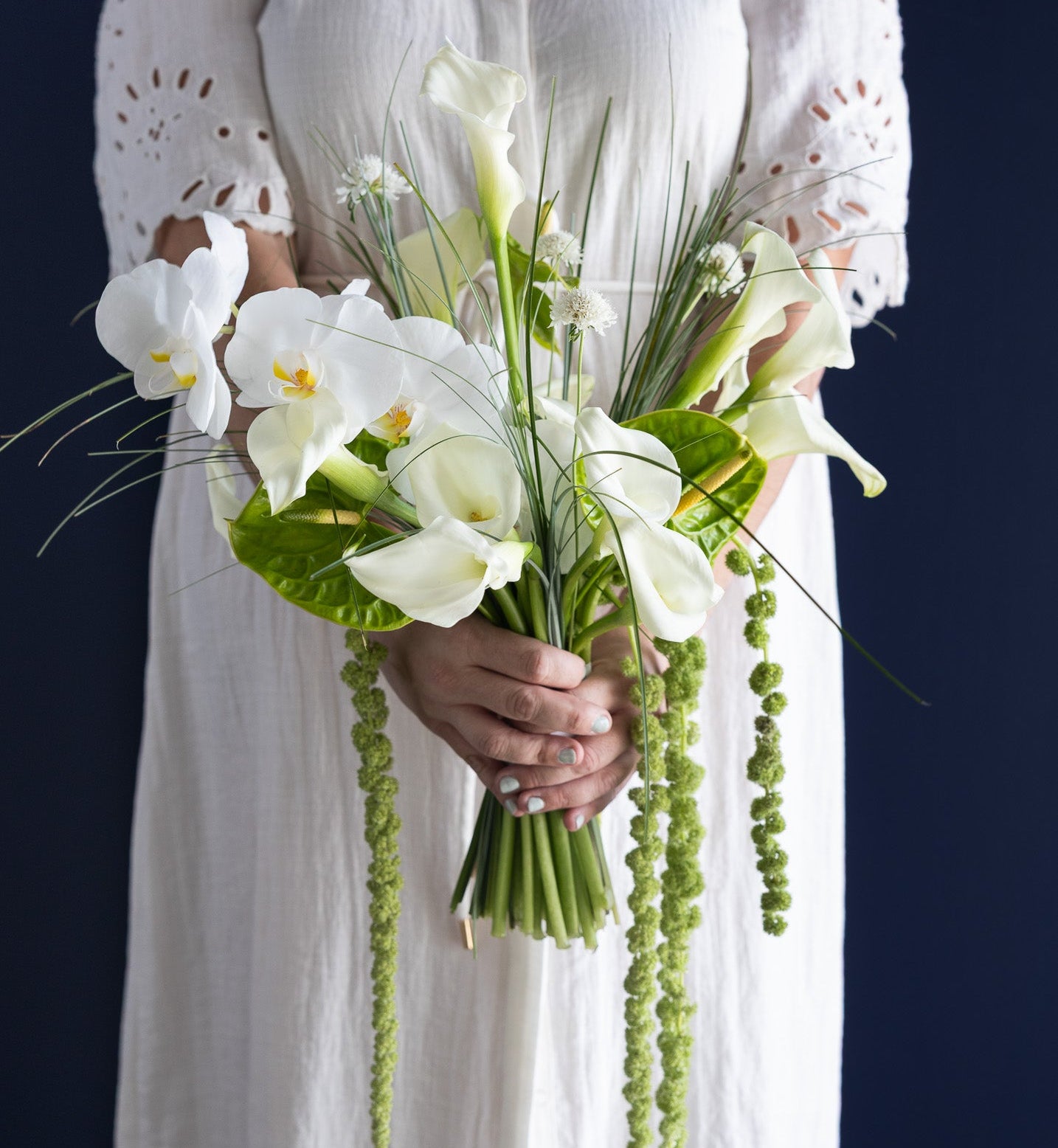 A person in a white eyelet dress holds a bouquet of white calla lilies, green foliage, and hanging green bead strands against a dark navy background.