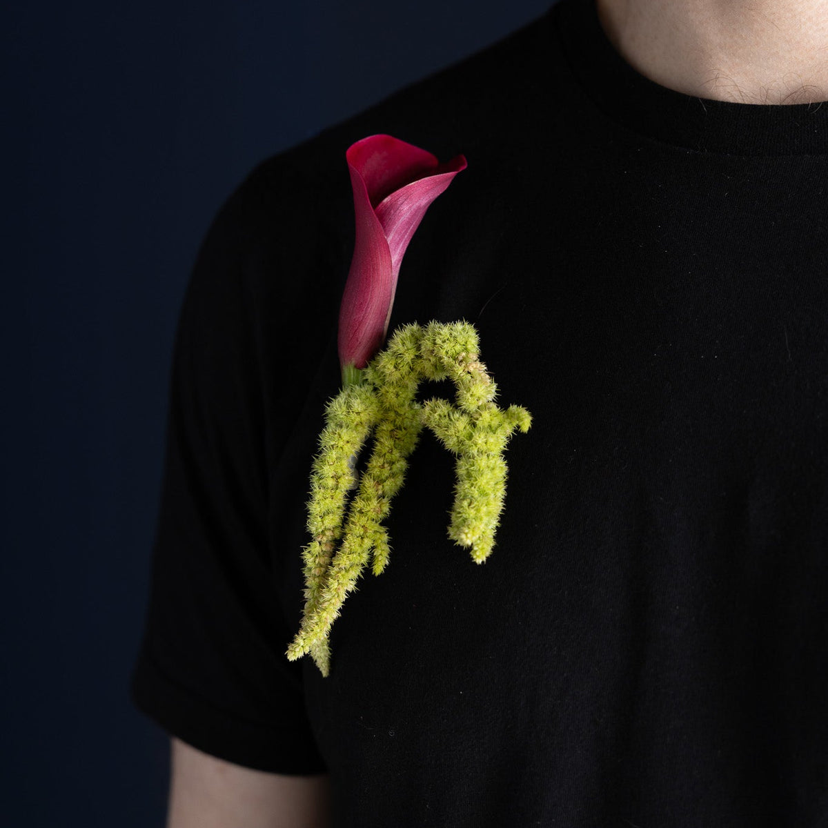 A close-up of a person wearing a black crew-neck t-shirt with a unique graphic print on the chest depicting a pink calla lily and a textured lime-green plant stem, set against a dark navy background.