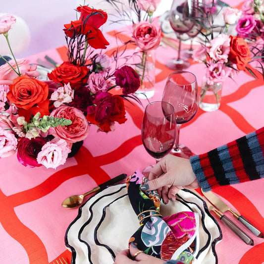Decorative table setting with flowers, glasses, and a colorful napkin on a pink and red patterned tablecloth.
