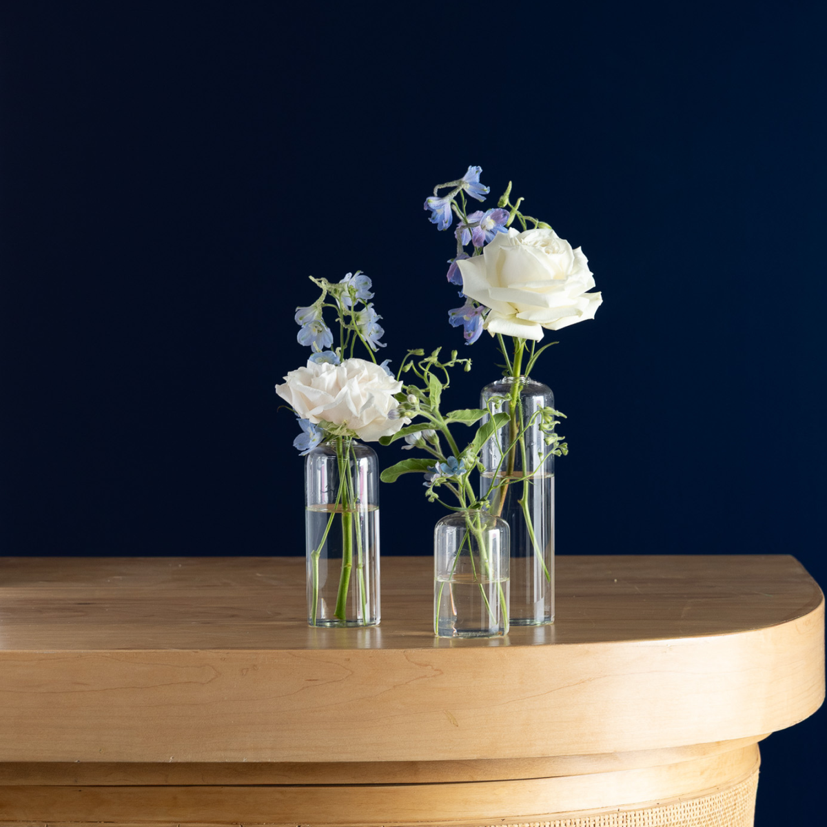 Three clear glass cylindrical vases with rounded tops, arranged on a light wood table against a dark navy blue background. The vases contain water with white roses and blue delphiniums.