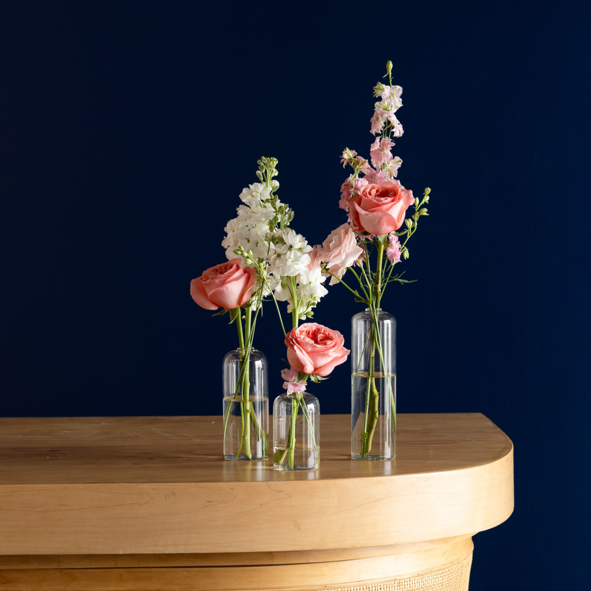 Three clear glass vases with different heights, arranged on a light wood console table against a dark navy blue background. The vases are filled with pink roses, white stock flowers, and pink delphiniums.