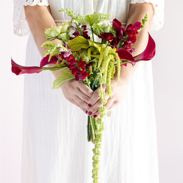 A person in a white dress holds a vibrant bouquet of flowers, including red calla lilies, green agapanthus, and burgundy snapdragons, against a clean white background.