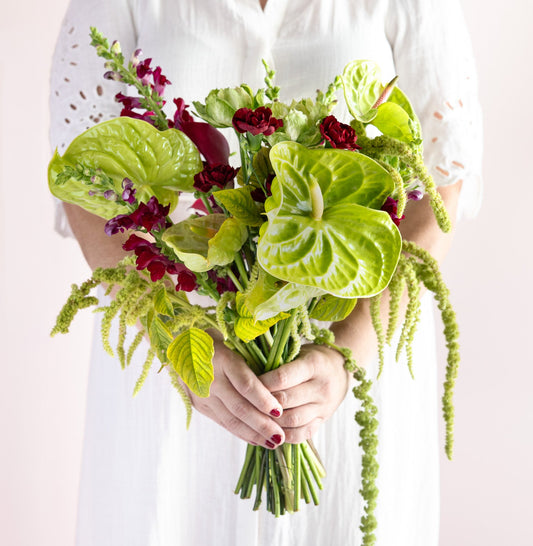 A person in a white blouse holds a lush bouquet of green anthuriums, burgundy roses, and green trailing spikes against a soft light pink background.
