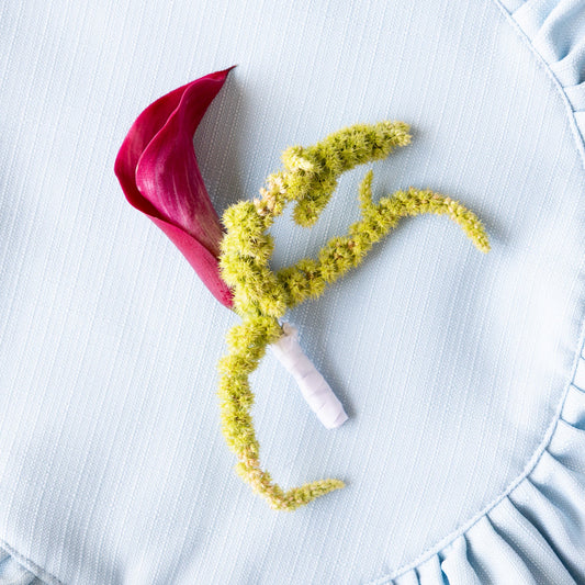 A floral boutonniere with a deep magenta calla lily and a sprig of yellow-green flowers, pinned to a light blue ruffled fabric background.