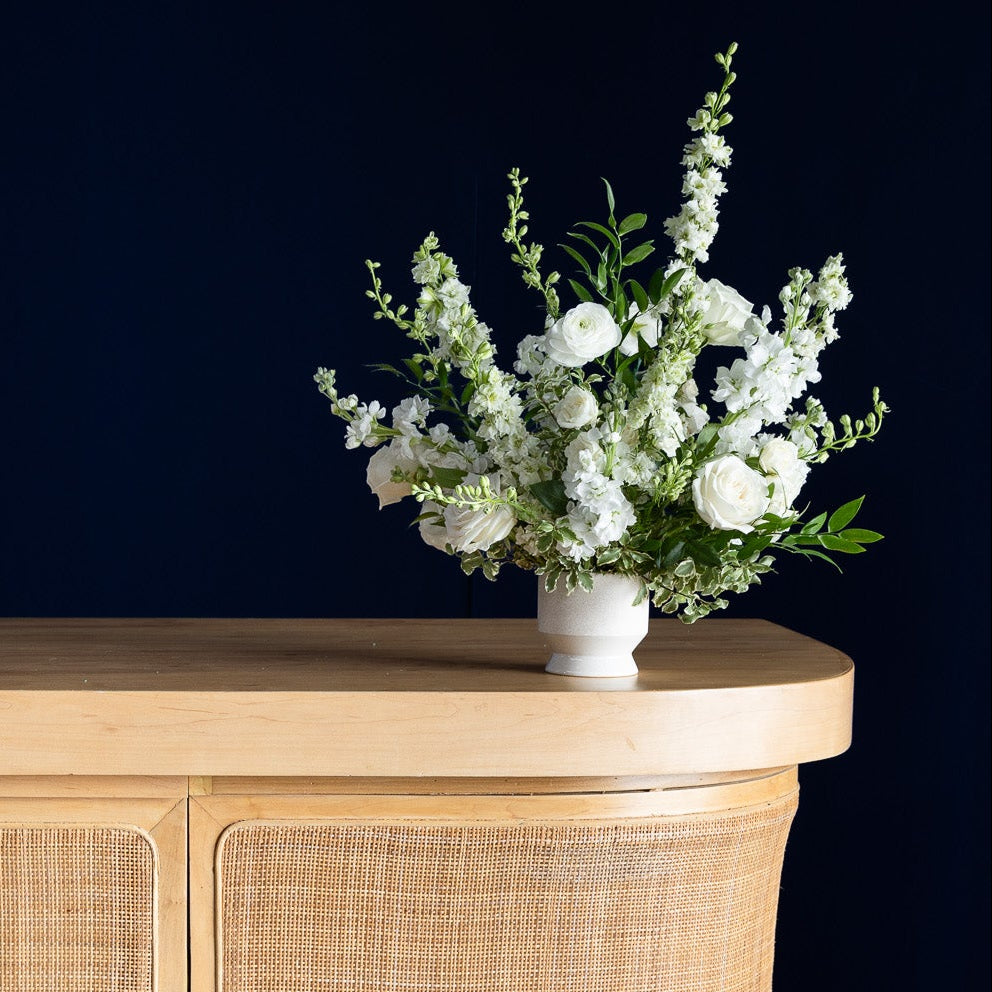 A curved wooden bar or console table with woven cane panels, styled with a white flower arrangement, wine glasses, and a small table lamp against a dark navy background.