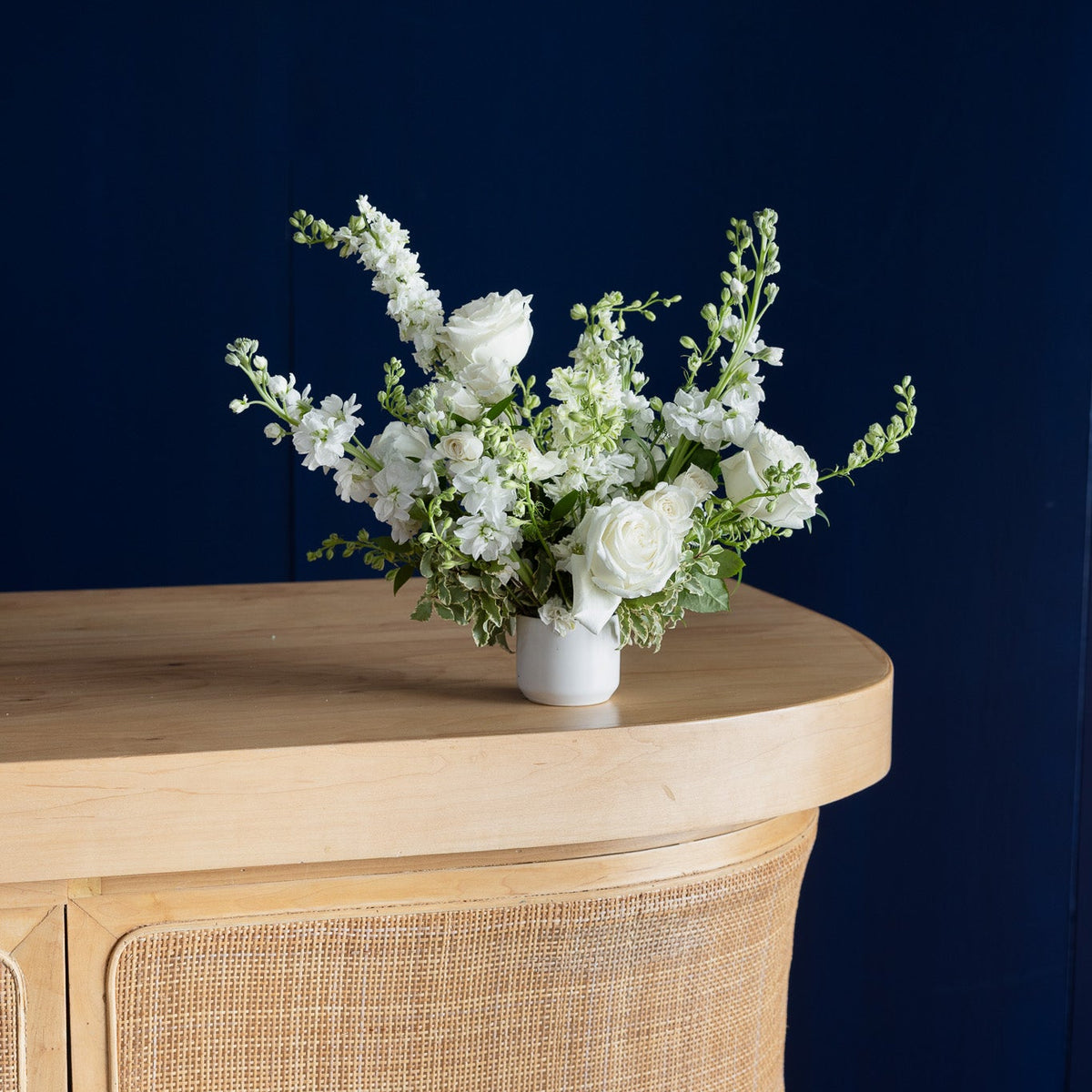 A white ceramic vase filled with a white rose and green foliage floral arrangement, displayed on a light wood console table against a dark navy blue background.