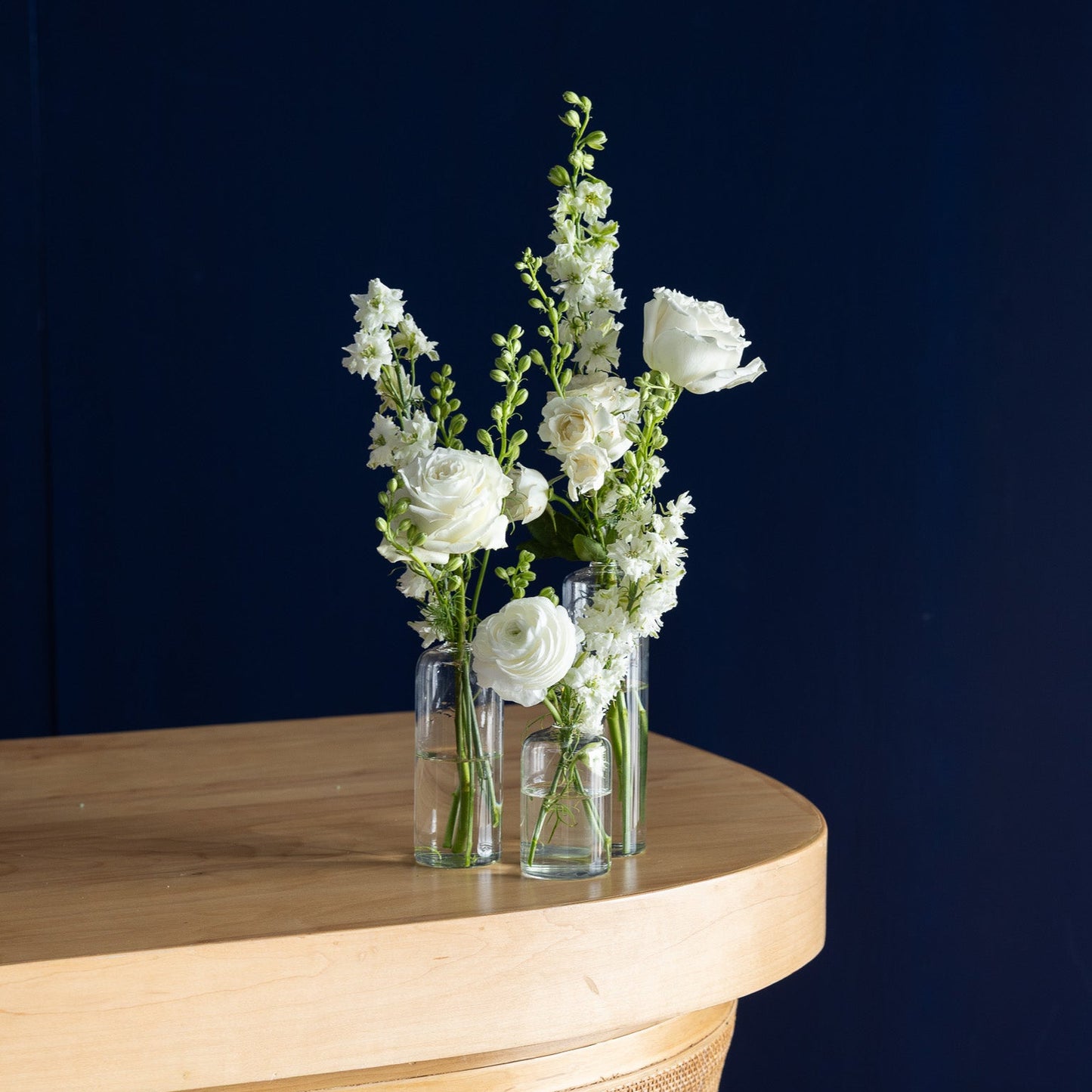 A clear glass vase holding white roses and green stems, placed on a curved light wood table against a dark navy blue background.