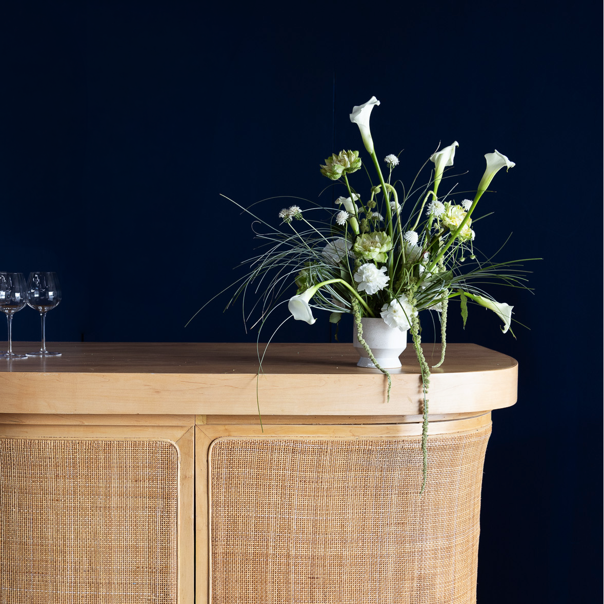 A curved wooden bar or console table with woven cane panels, styled with a small lamp, wine glasses, and a white floral arrangement against a dark navy background.
