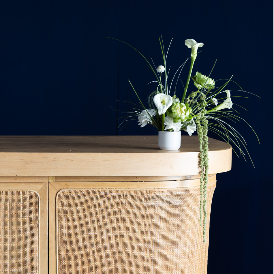A curved wooden bar cart with woven cane panels, styled with a small table lamp, wine glasses, and a floral arrangement against a dark navy background.