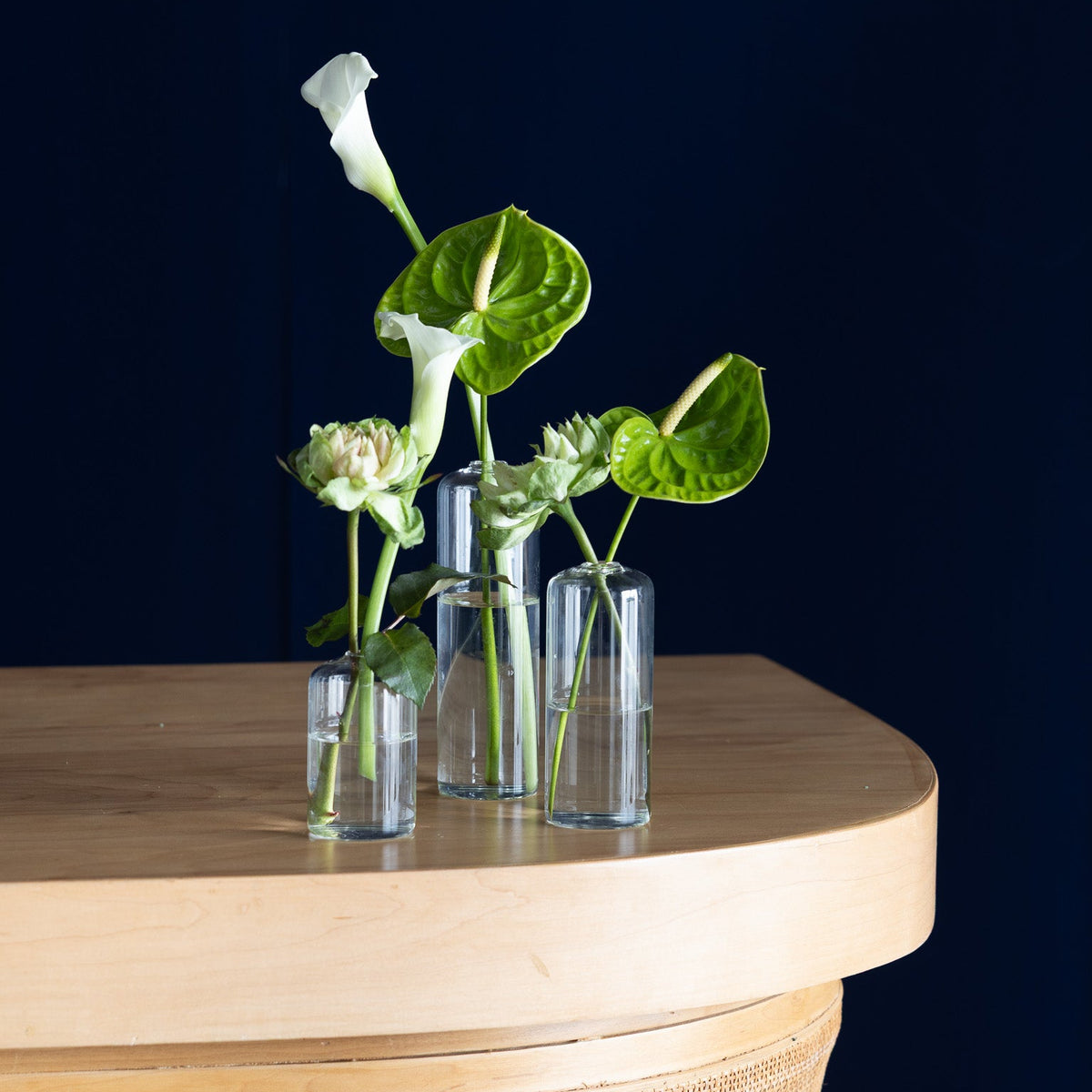 Three clear glass cylinder vases with green stems, white calla lilies, and green anthurium leaves arranged on a light wood table against a dark navy blue background.