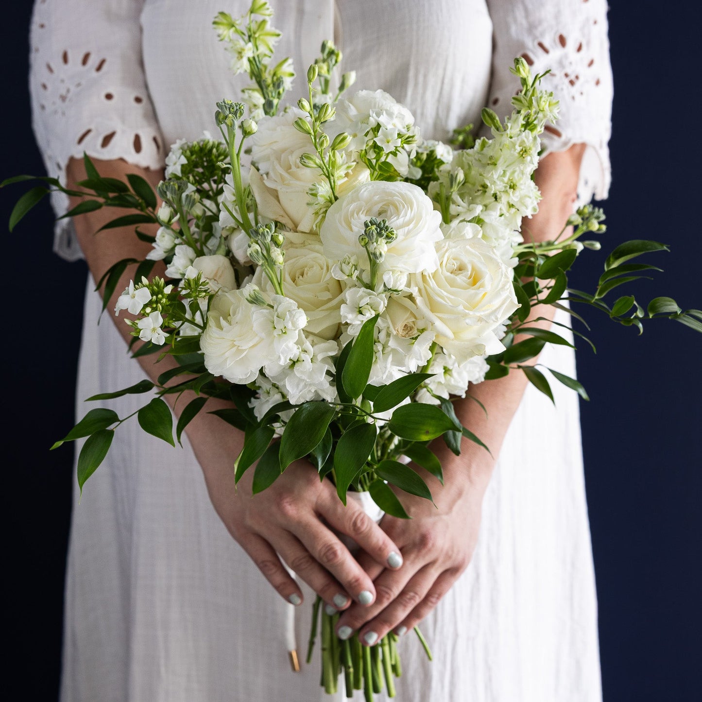A person in a white dress holding a bouquet of white flowers and green foliage against a dark navy background.