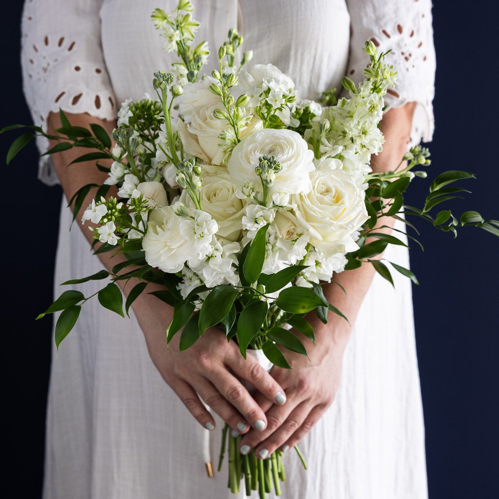 A person in a white dress holding a bouquet of white flowers and green foliage against a dark navy background.