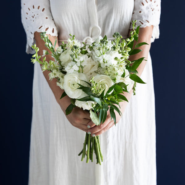 A person wearing a white eyelet dress holds a bouquet of white flowers and green foliage against a dark navy background.